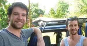 2 men in front of blue pick up with surfboard on top